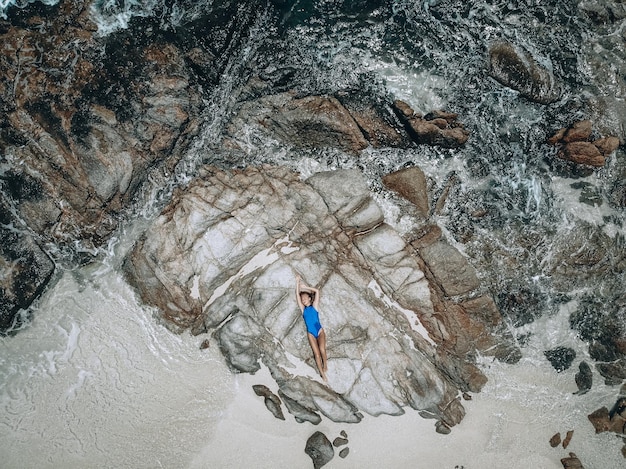 Dall'alto foto di una bella giovane donna con un costume da bagno blu sdraiato sulla roccia durante l'alta marea. Concetto di vacanza estiva