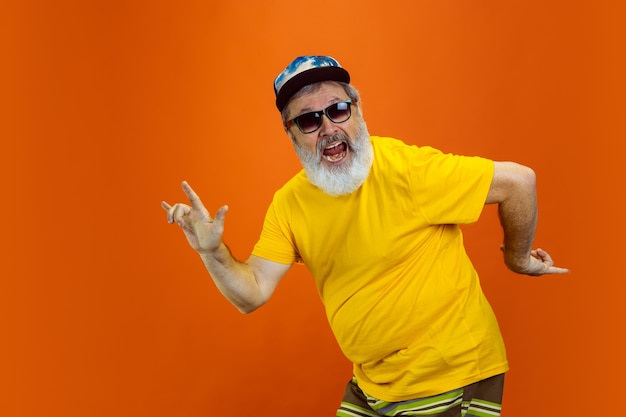 Danza pazzesca. Ritratto di un uomo anziano hipster che utilizza dispositivi, gadget isolati sulla parete arancione dello studio