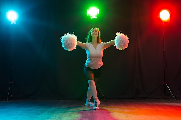 Danza, sport, bellezza e concetto di persone - la giovane ragazza cheerleader nell'oscurità mostra pon pon e sorride.