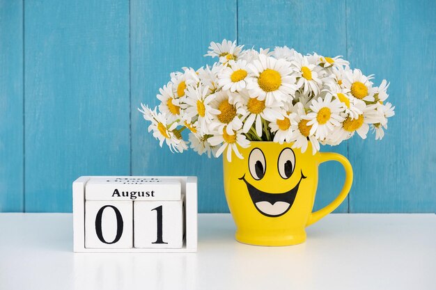 Data del 1° agosto sul calendario e bouquet di camomilla in tazza gialla con faccia buffa contro il muro blu Ciao concetto del mese estivo di agosto