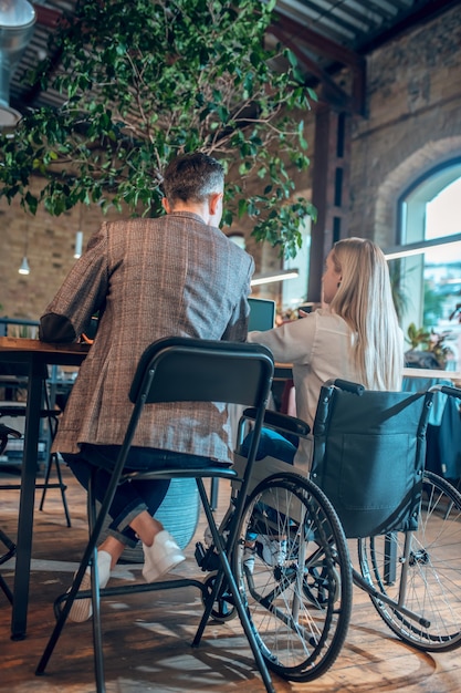 Data. Donna con lunghi capelli biondi in sedia a rotelle e uomo in giacca e jeans seduto al tavolo con le spalle alla telecamera