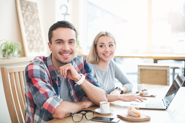 Data in caffè. Giovani coppie in caffè con interni eleganti. Studenti che bevono deliziose bevande al caffè. Chiacchierano, usano il laptop e sorridono. Uomo che guarda la telecamera