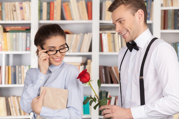 Data in libreria. Allegro giovane nerd che dà una rosa rossa a una bellissima giovane donna con gli occhiali mentre si trova in biblioteca