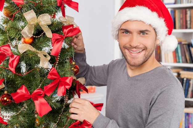 Decorare un albero di Natale. Giovane allegro che decora un albero di Natale e sorride alla macchina fotografica