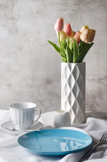 Decorazione della tavola di primavera mattutina. Piatto ceramico vuoto, tazza, tulipani freschi in vaso sul fondo bianco pulito della tovaglia. Concetto di cena o colazione,