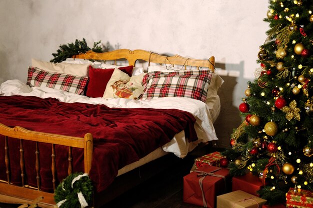 Decorazioni natalizie per la camera da letto Un grande letto in legno con copriletto rosso e cuscini a scacchi e un albero di Natale in un angolo della stanza