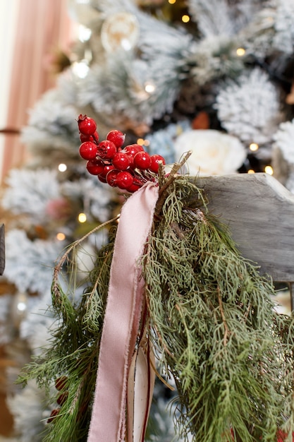 Decorazioni per la casa di Natale o Capodanno sulla sedia, nastri, bacche e rami, albero di Natale sullo sfondo. Dettagli della celebrazione delle vacanze invernali