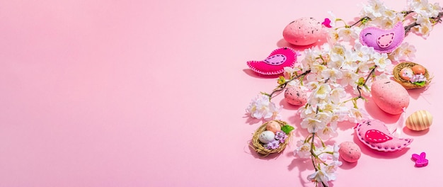 Delicata composizione pasquale con fiori di ciliegio e uccelli in feltro fatti a mano Uova decorative e nidificano simpatici conigli Formato banner di sfondo rosa chiaro scuro ombra dura