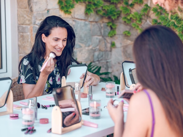 Deliziosa bruna e anonima signora sfocata sedute insieme a tavolo con i cosmetici e applicando il trucco contro il muro dell'edificio alla luce del giorno