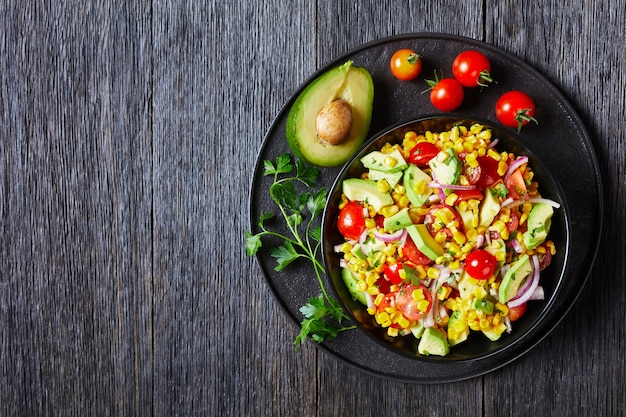 Deliziosa insalata di mais dolce con avocado, fette di cipolla rossa e pomodori in una ciotola nera su un tavolo di legno scuro, cucina messicana, vista orizzontale dall'alto, piatto, spazio libero