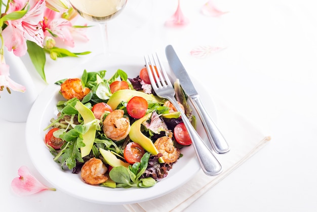 Deliziosa insalata fresca con gamberi, verdure, cetrioli, avocado e pomodori con vino bianco e fiori su superficie chiara