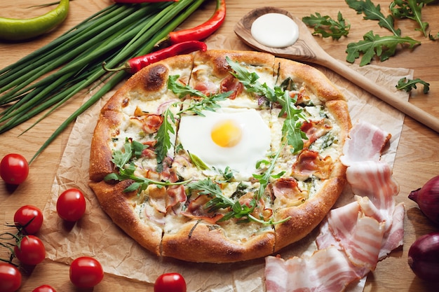 Deliziosa pizza con pancetta, uova e rucola servita su un tavolo di legno circondato da ingredienti freschi