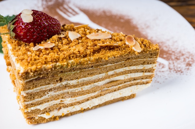 Deliziosa torta di mandorle a strati con un ripieno cremoso di noci sormontato da una fragola fresca e menta servita su un piatto con sagome decorative di polvere di cioccolato di una forchetta e un cucchiaio, vista ravvicinata