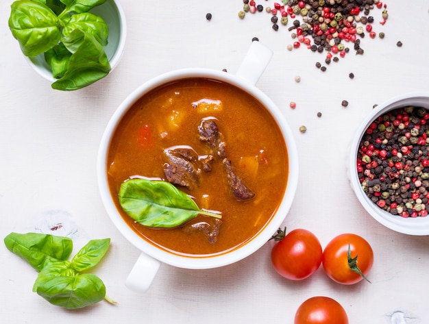 Deliziosa zuppa di pomodoro con carne su un tavolo in legno rustico bianco con pomodorini freschi, foglie di basilico e pepe secco. Ingredienti per la zuppa. Vista dall'alto