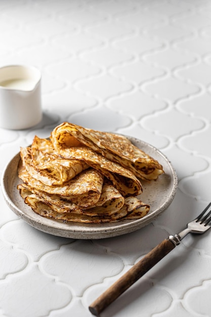 Deliziose crepes fatte in casa su piatto di ceramica con forchetta e salsiera su sfondo piastrellato bianco per maslenitsa