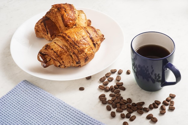 Deliziosi croissant croccanti appetitosi caffè caldo nella tazza blu e asciugatutto sulla piastra sul tavolo di marmo