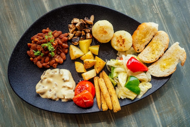 Deliziosi fagioli rossi sul pane con patate e pomodoro Pannocchie di mais fritte con funghi su piatto nero pasto biologico vegano