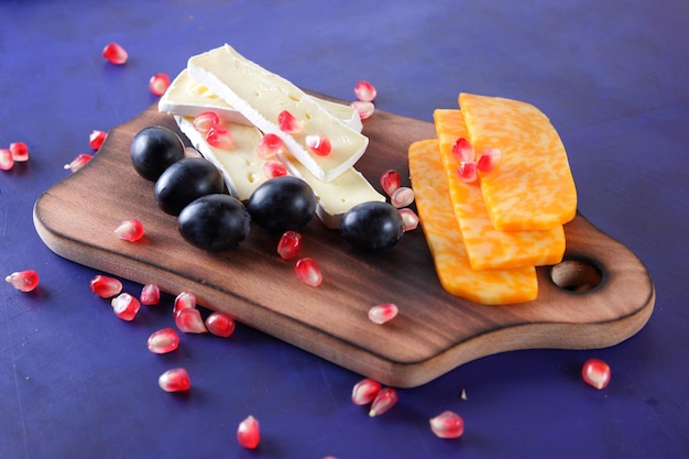 Deliziosi formaggi su una tavola di legno closeup Camembert e fette di formaggio a pasta dura gialla con semi di melograno e uva scura su sfondo blu
