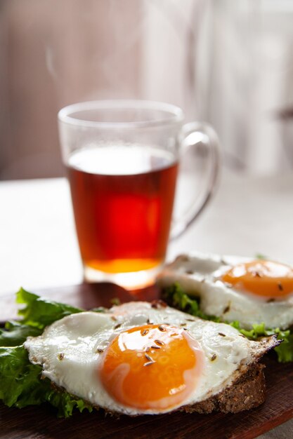 Deliziosi toast con uova fritte e foglie di lattuga per colazione posti sul tavolo vicino a una tazza di tè caldo al mattino