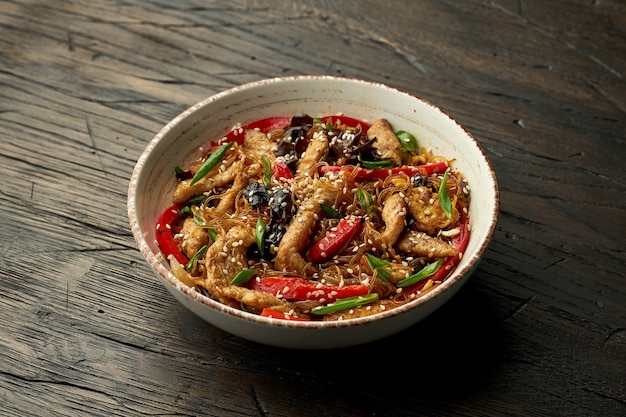 Delizioso cibo asiatico di strada - tagliatelle funchose con pollo, coriandolo, verdure e frittata in una ciotola bianca su una superficie di legno