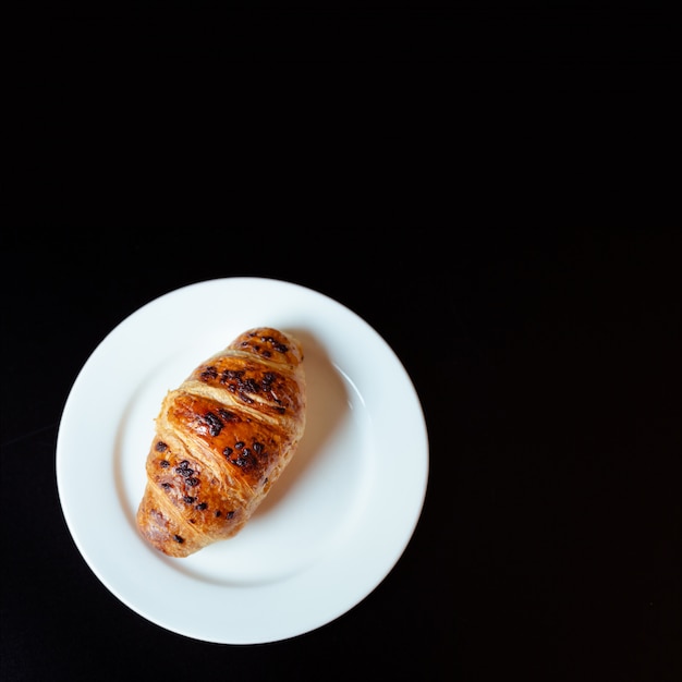 Delizioso croissant dessert-croccante con gocce di cioccolato su un piatto bianco si erge su una superficie nera.