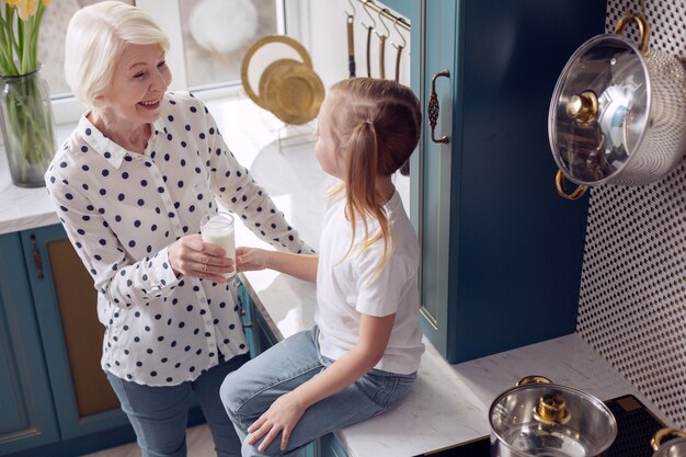 Delizioso latte. La vista dall'alto di un'affascinante bambina seduta sul bancone della cucina e prendendo un bicchiere di latte dalle mani della sua amata nonna