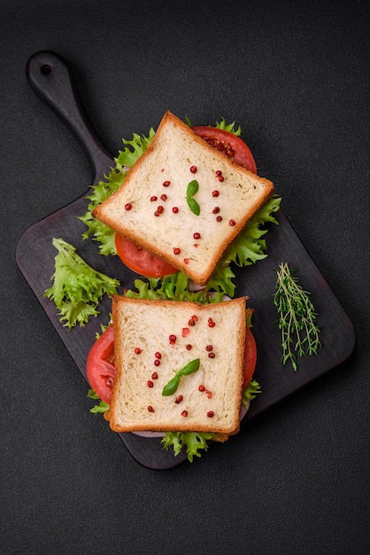 Delizioso panino con toast prosciutto pomodori formaggio e lattuga con sale spezie ed erbe aromatiche su uno sfondo di cemento scuro