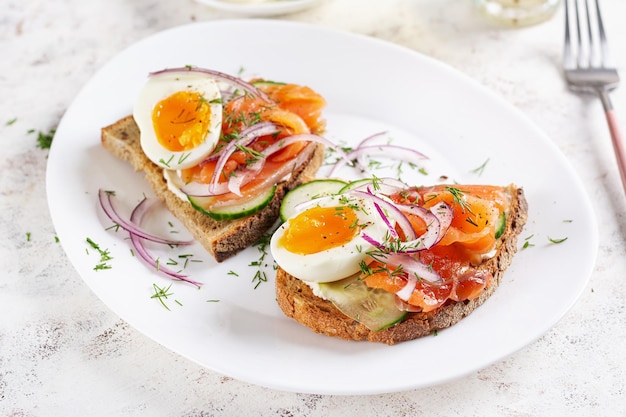 Delizioso toast con salmone, cetriolo bollito, uova e formaggio a crema su un piatto bianco.