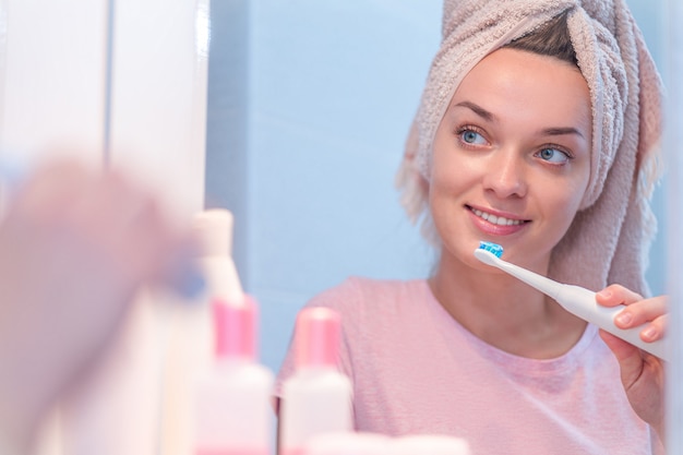 Denti di spazzola felici della donna in buona salute facendo uso dello spazzolino da denti elettrico ultrasonico nel bagno a casa