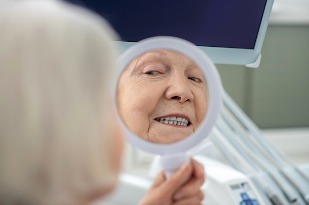 Denti dopo il restauro. Senior donna seduta presso l'ufficio dei dentisti e guardando allo specchio dopo il restauro dei denti
