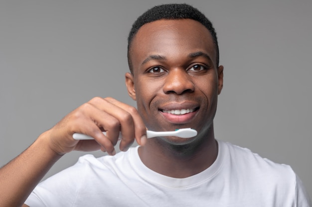 Denti sani. Afroamericano attraente allegro con lo spazzolino da denti vicino alla bocca sorridente su fondo grigio chiaro