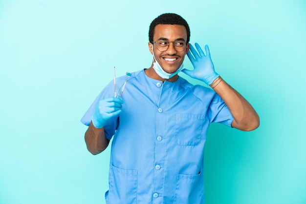 Dentista afroamericano che tiene gli strumenti su sfondo blu isolato ascoltando qualcosa mettendo la mano sull'orecchio