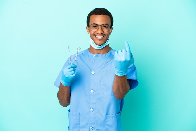 Dentista afroamericano che tiene gli strumenti su sfondo blu isolato facendo un gesto imminente