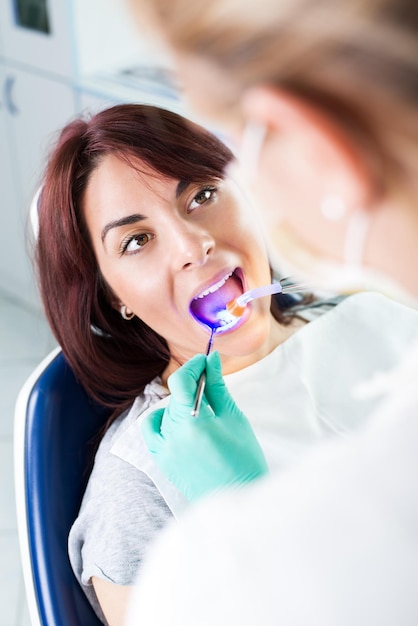 Dentista con lampada UV a polimerizzazione dentale che rifinisce il dente di riparazione sul paziente femminile in ufficio. Focus selettivo, focus sul paziente.