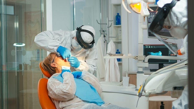 Dentista con viso shiled che tratta i denti a una paziente anziana in clinica odontoiatrica durante il covid-19. Medico e infermiere che lavorano indossando tuta, tuta protettiva, maschera, guanti in ufficio stomatologico.