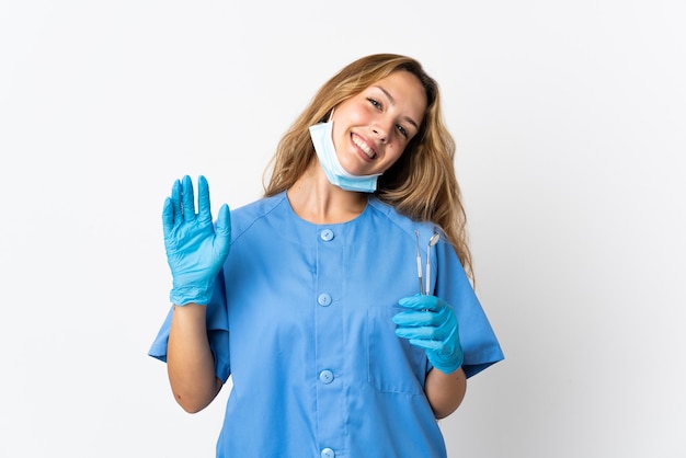 Dentista della donna che tiene gli strumenti isolati sulla parete bianca che saluta con la mano con l'espressione felice