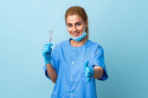 Dentista della giovane donna che tiene gli strumenti sopra l'handshaking blu isolato della parete dopo un buon affare