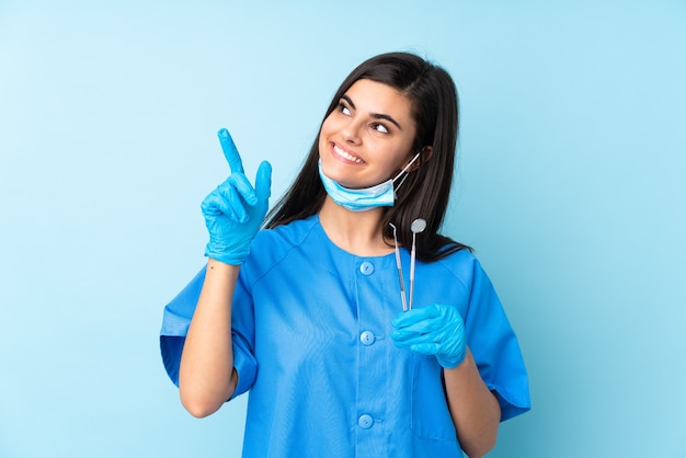 Dentista della giovane donna che tiene gli strumenti sopra la parete blu isolata che indica con il dito indice una grande idea