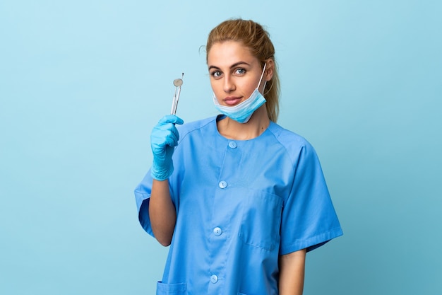 Dentista della giovane donna che tiene gli strumenti sopra la parete blu isolata mantenendo le braccia incrociate