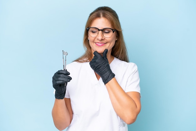 Dentista di mezza età che tiene gli strumenti su sfondo blu isolato guardando di lato e sorridente