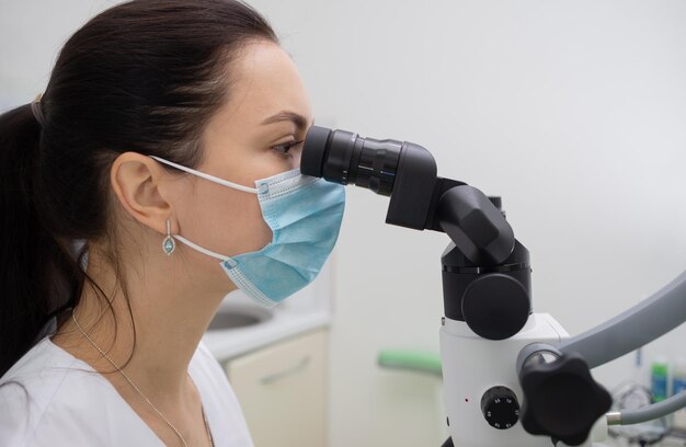 Dentista donna bruna in camice bianco e maschera protettiva medica lavora con un microscopio moderno in ufficio Operazioni nella clinica dentale