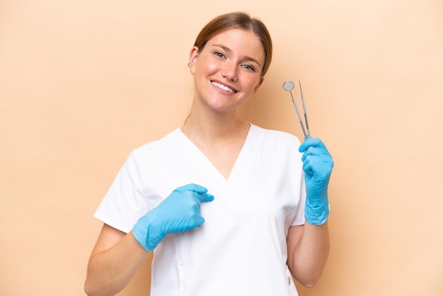 Dentista donna caucasica che tiene gli strumenti isolati su sfondo beige con espressione facciale a sorpresa