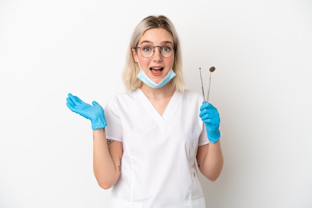 Dentista donna caucasica che tiene gli strumenti isolati su sfondo bianco con espressione facciale scioccata