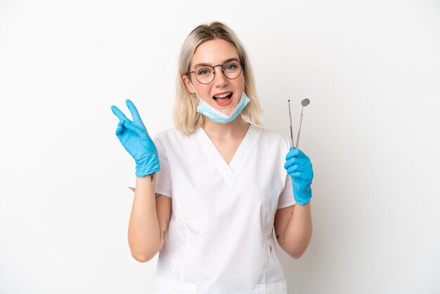 Dentista donna caucasica che tiene strumenti isolati su sfondo bianco sorridente e mostrando il segno di vittoria