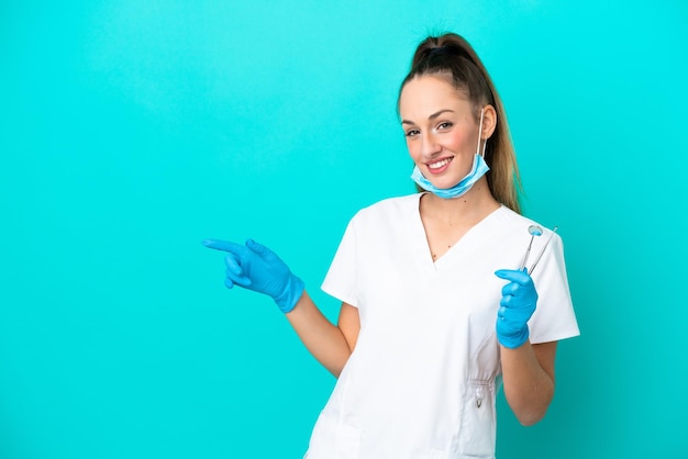 Dentista donna caucasica che tiene strumenti isolati su sfondo blu che punta il dito di lato