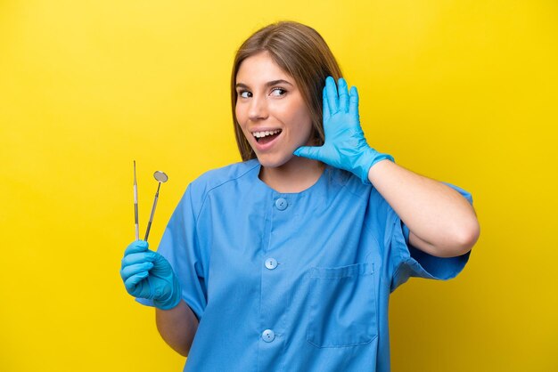 Dentista donna caucasica che tiene strumenti isolati su sfondo giallo ascoltando qualcosa mettendo la mano sull'orecchio
