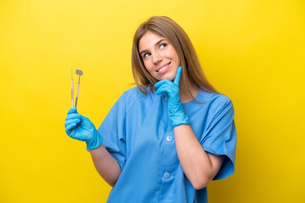 Dentista donna caucasica che tiene strumenti isolati su sfondo giallo pensando a un'idea mentre si guarda in alto