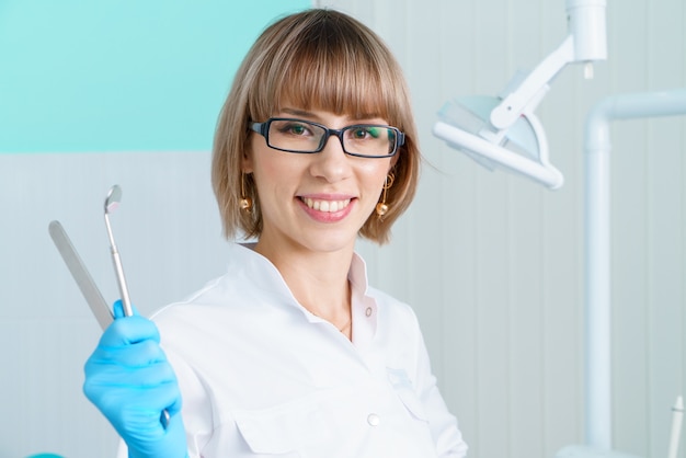 Dentista donna che tiene gli strumenti sullo sfondo dello studio dentistico in camice bianco e occhiali sorride...