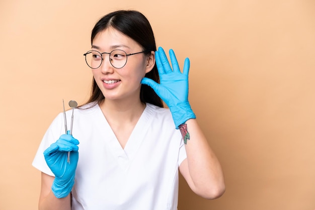 Dentista donna cinese che tiene strumenti isolati su sfondo beige ascoltando qualcosa mettendo la mano sull'orecchio