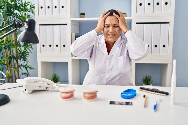 Dentista donna ispanica matura che lavora presso la clinica che soffre di mal di testa disperata e stressata a causa del dolore e dell'emicrania. mani sulla testa.